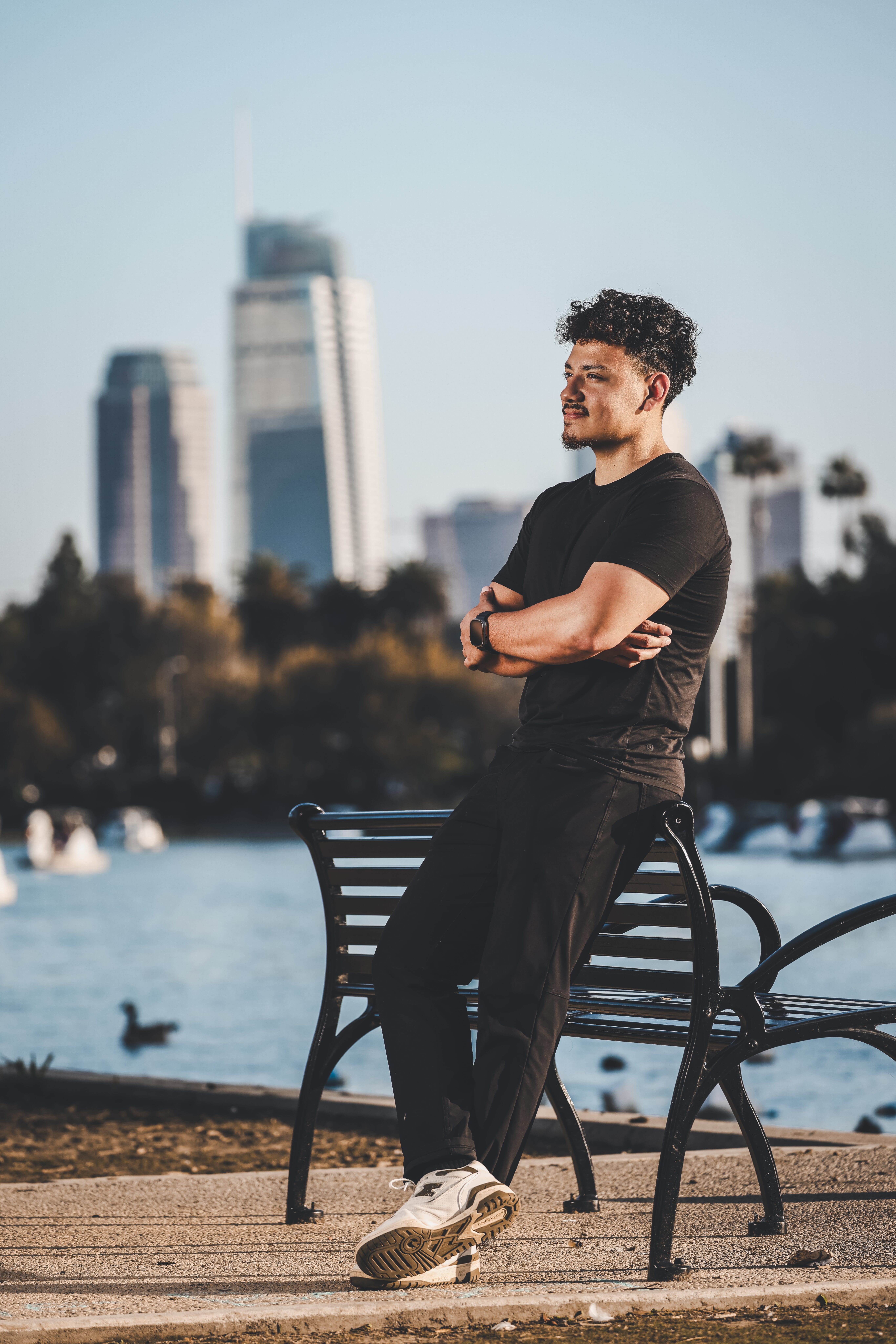 Osiel Campos Jr. standing confidently by a lakeside bench with the Los Angeles skyline in the background, representing his roots and reputation in LA and OC.