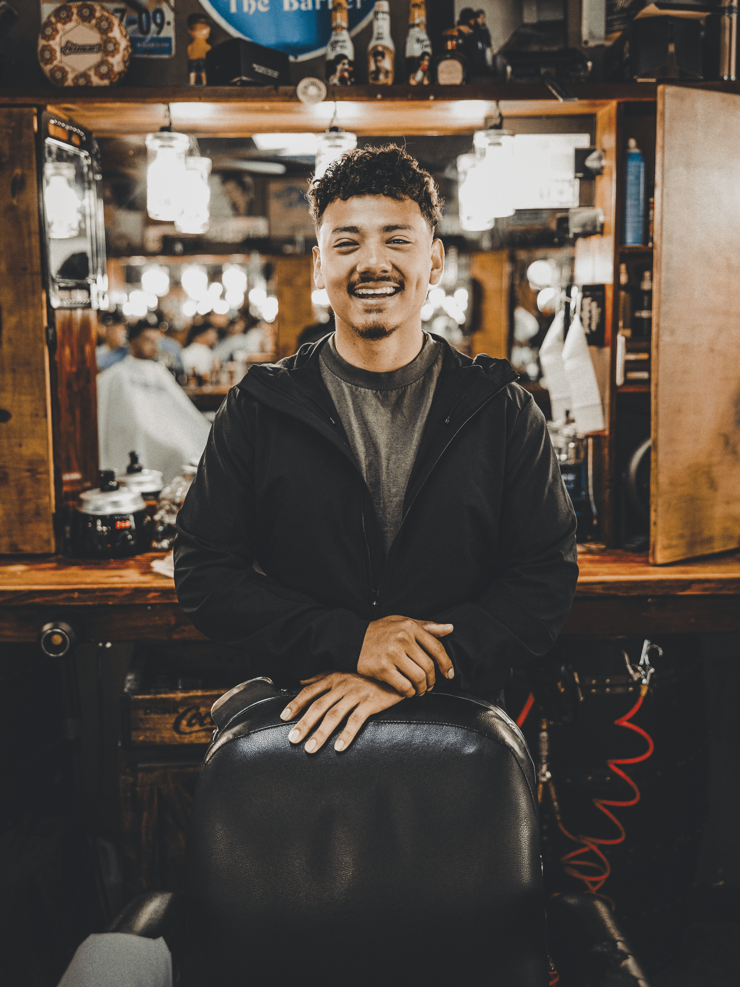 Barber Osiel smiling while seated in front of a mirror at the barbershop, showcasing experience and personal connection with clients.